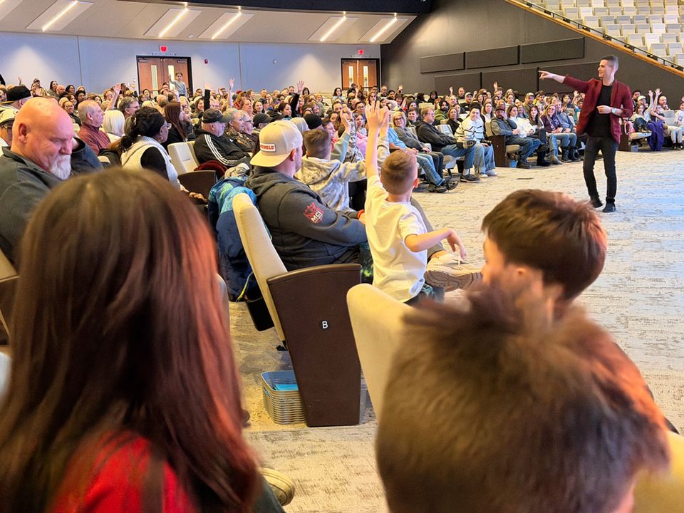 A church auditorium full of families watching the gospel magic show, children with hands raised to volunteer